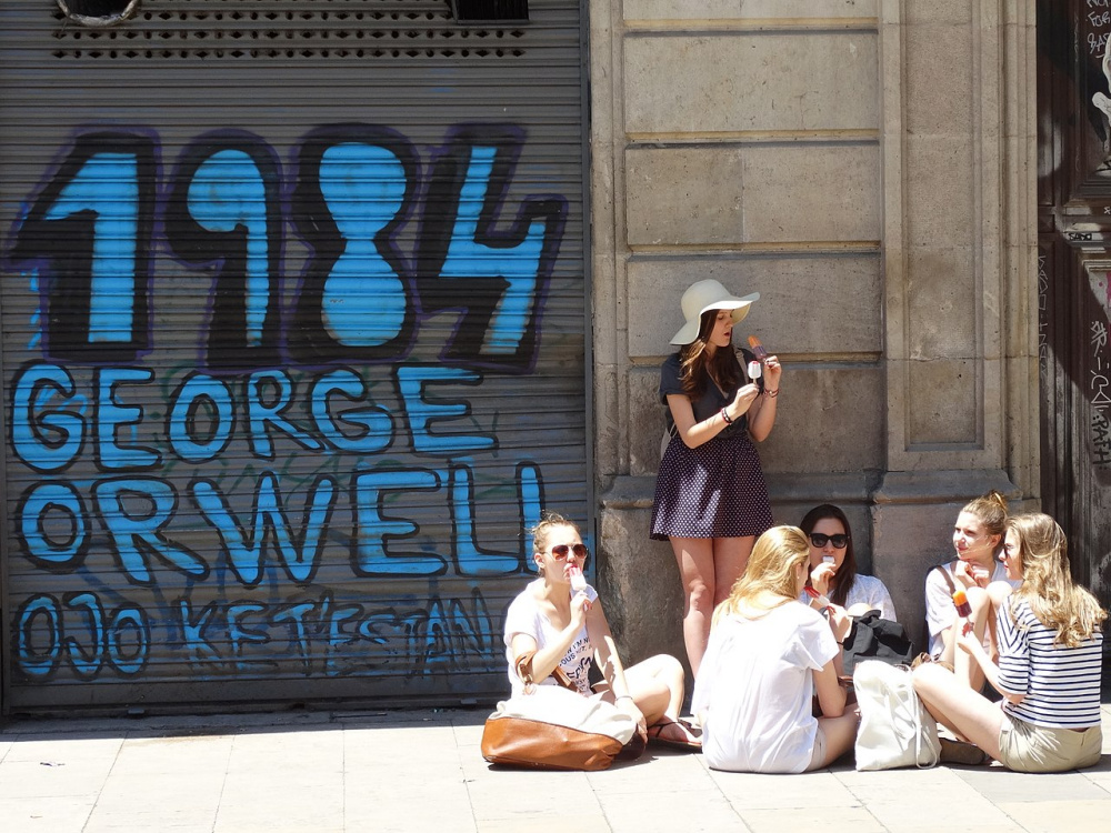 Graffiti in the Plaça de George Orwell, Gothic Quarter, Barcelona. Painted on a metal shutter are the words '1984 George Orwell' in large blue letters, while a group of young people eat ice cream in front of the wall.