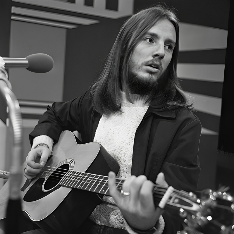 Dewey Bunnell playing guitar on the TopPop TV show in 1972. He is wearing a white jumper and a dark jacket.