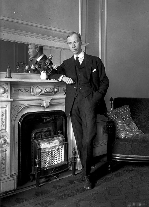 Sergei Prokofiev standing beside a fireplace, circa 1918, in a formal suit, with a faint reflection in the mirror behind him